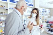 Help from a pharmacist. A female pharmacist with glasses holds medicine boxes and instructions for use. She looks at the man who is explaining something else. They both wear protective masks