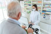 Buying and selling prescription drugs and pharmacist advice. An adult female pharmacist standing behind the counter and selling drugs to a mature man. She is wearing a protective mask